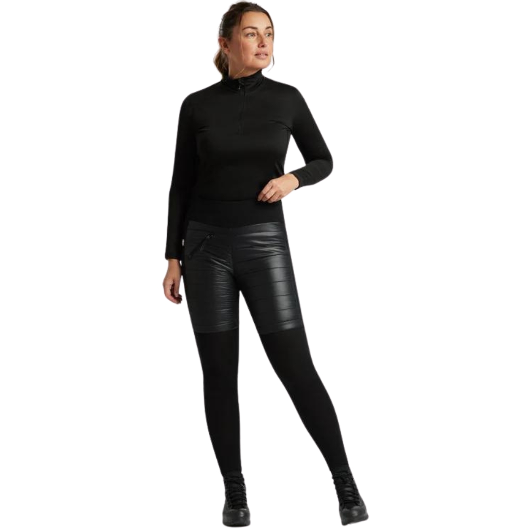 Une femme est debout et porte une tenue noire. Elle porte un haut noir à manches longues et un pantalon noir slim avec une texture semblable à du cuir sur la partie avant. Elle a une main sur la hanche et regarde sur le côté. Elle porte également des chaussures noires et a les cheveux attachés en arrière.