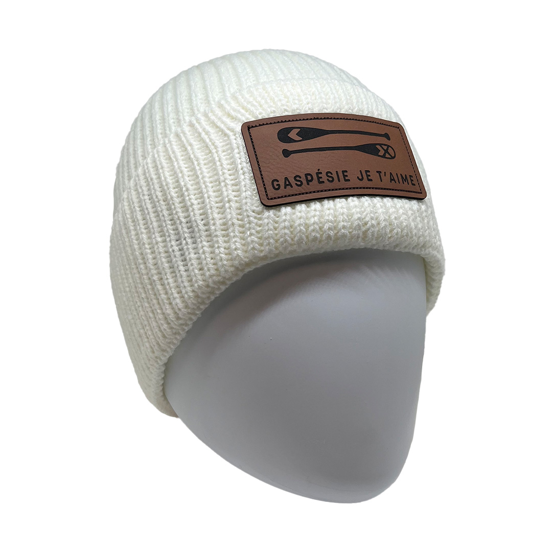 Un bonnet tricoté blanc avec un patch marron sur le devant qui présente un design minimaliste de skis croisés et le texte « GASPÉSIE JE T'AIME! » en noir. Le bonnet est présenté sur une tête de mannequin unie et blanche.