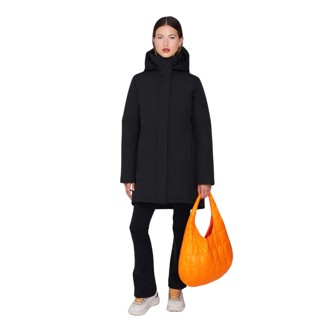 Une personne portant un manteau à capuche noir et un pantalon noir se tient debout sur un fond blanc, tenant un grand sac orange matelassé. Elle porte des baskets blanches et a une expression neutre.