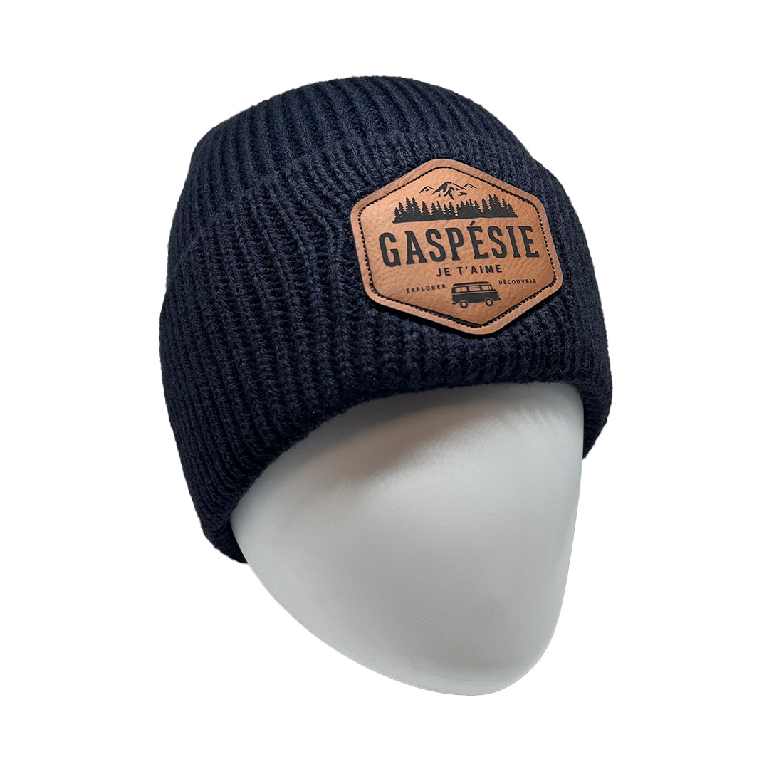 Un bonnet bleu foncé avec un patch marron affichant « Gaspésie Je T'aime » et une illustration de montagnes, d'arbres et d'un camping-car d'époque. Le bonnet est présenté sur une tête de mannequin blanche.